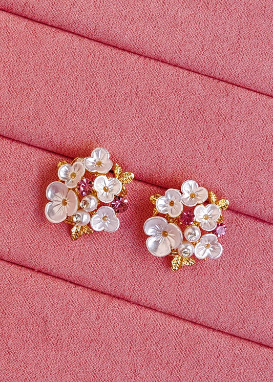 A pair of floral stud earrings with white mother-of-pearl flowers, pink accents, and gold details, displayed on a textured pink fabric background.