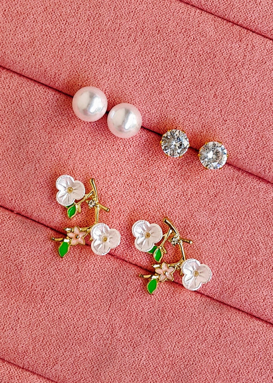 Two pairs of stud earrings arranged on a pink textured fabric background. One pair features smooth white pearls, and the other pair features small white flowers with green leaves and gold-tone metal settings.
