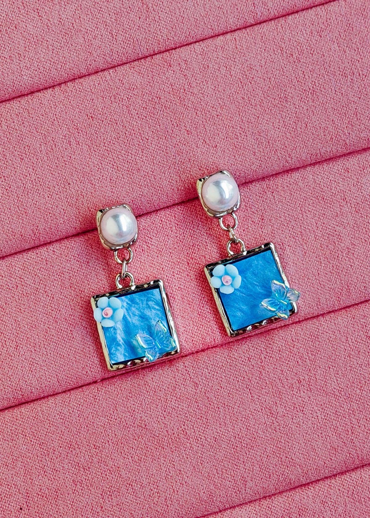 A pair of drop earrings with white pearl-like studs and square blue floral charms in silver-toned settings, displayed on a pink textured surface.