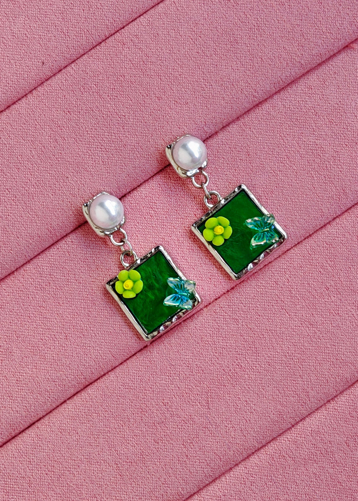A pair of silver dangle earrings with green square enamel panels, yellow flower charms, and small white pearl-like beads at the top, displayed on a pink textured background.