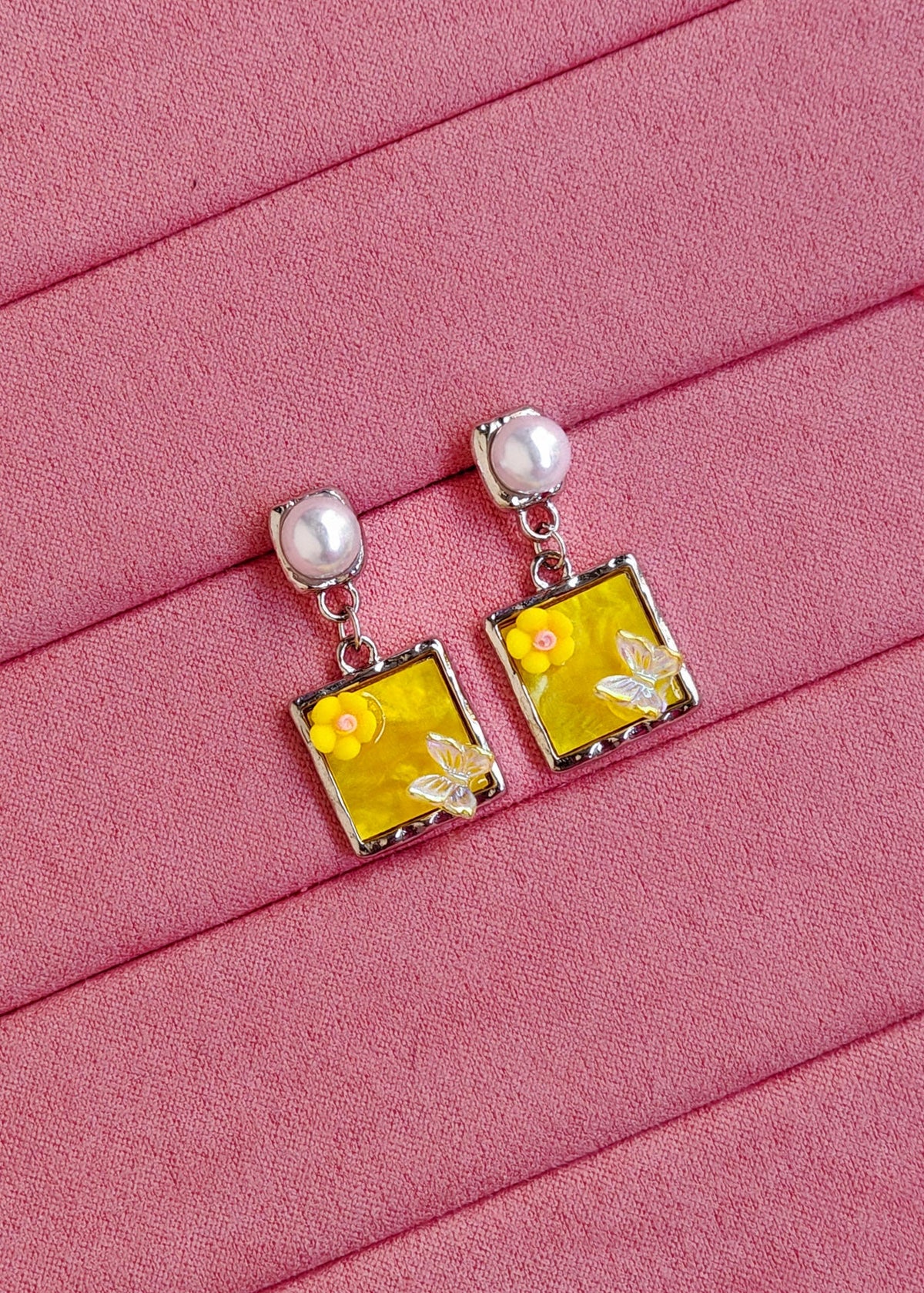 A pair of silver-tone dangle earrings with round white pearl studs and square yellow charms featuring a small yellow flower and a white bee, displayed on a pink textured fabric background.