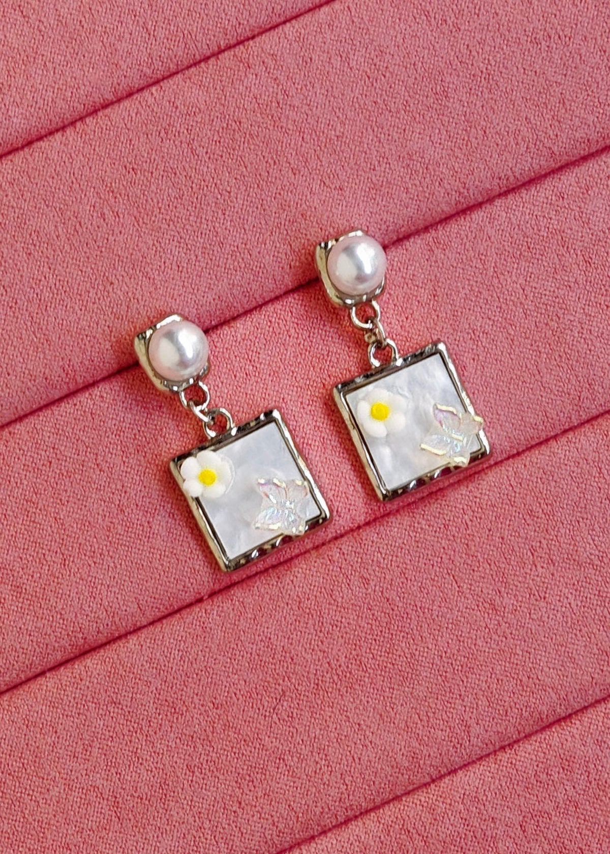 A pair of silver-toned square dangle earrings with white faux-pearl centers, yellow flower accents, and tiny clear rhinestones, displayed on a pink ribbed fabric background.