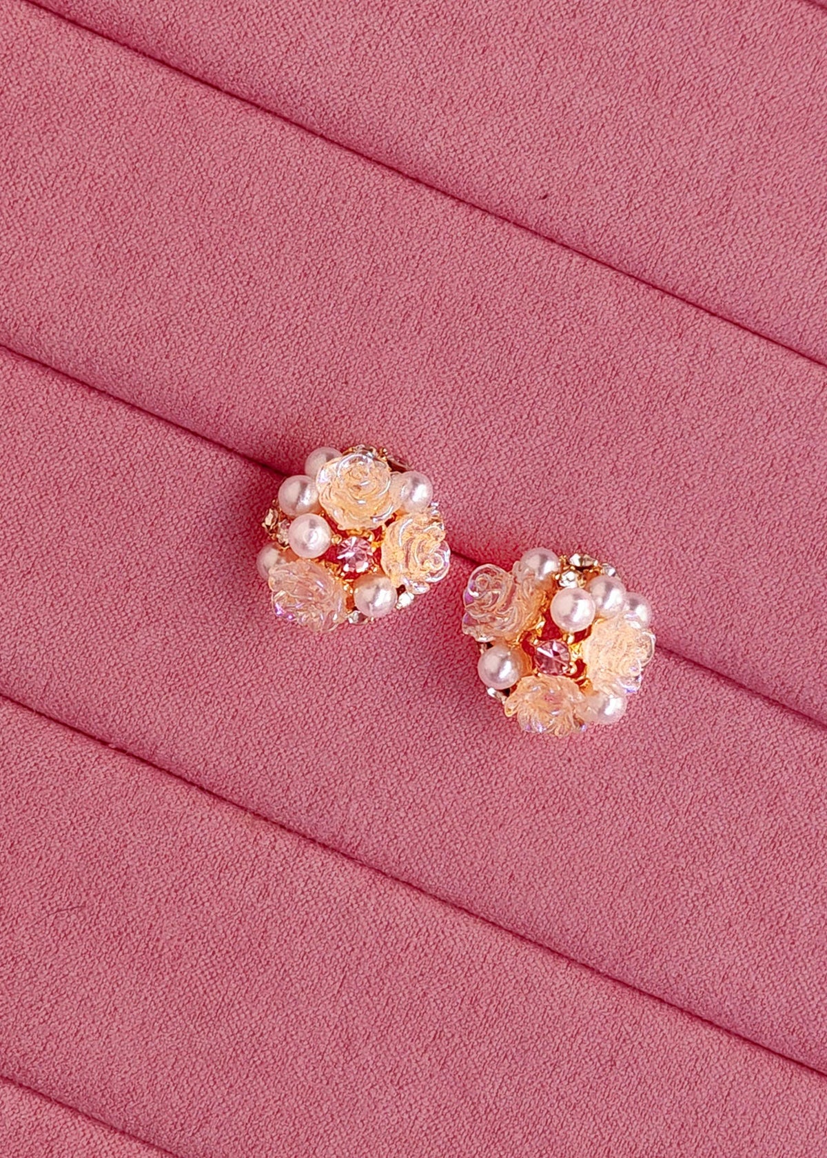 A pair of rose gold floral stud earrings decorated with faux pearls and clear rhinestones, displayed on a textured pink fabric background.
