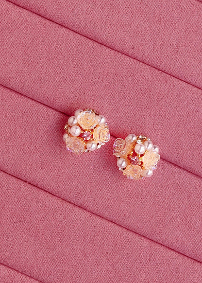 A pair of rose gold floral stud earrings decorated with faux pearls and clear rhinestones, displayed on a textured pink fabric background.