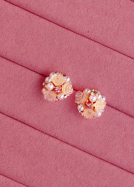A pair of rose gold floral stud earrings decorated with faux pearls and clear rhinestones, displayed on a textured pink fabric background.