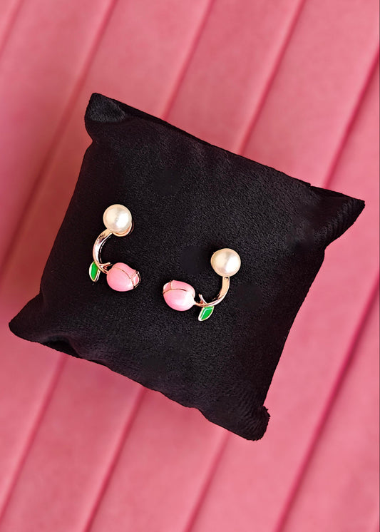 A pair of gold-toned belly button rings decorated with pink cherry charms and small green leaves, displayed on a black fabric pillow against a pink background.