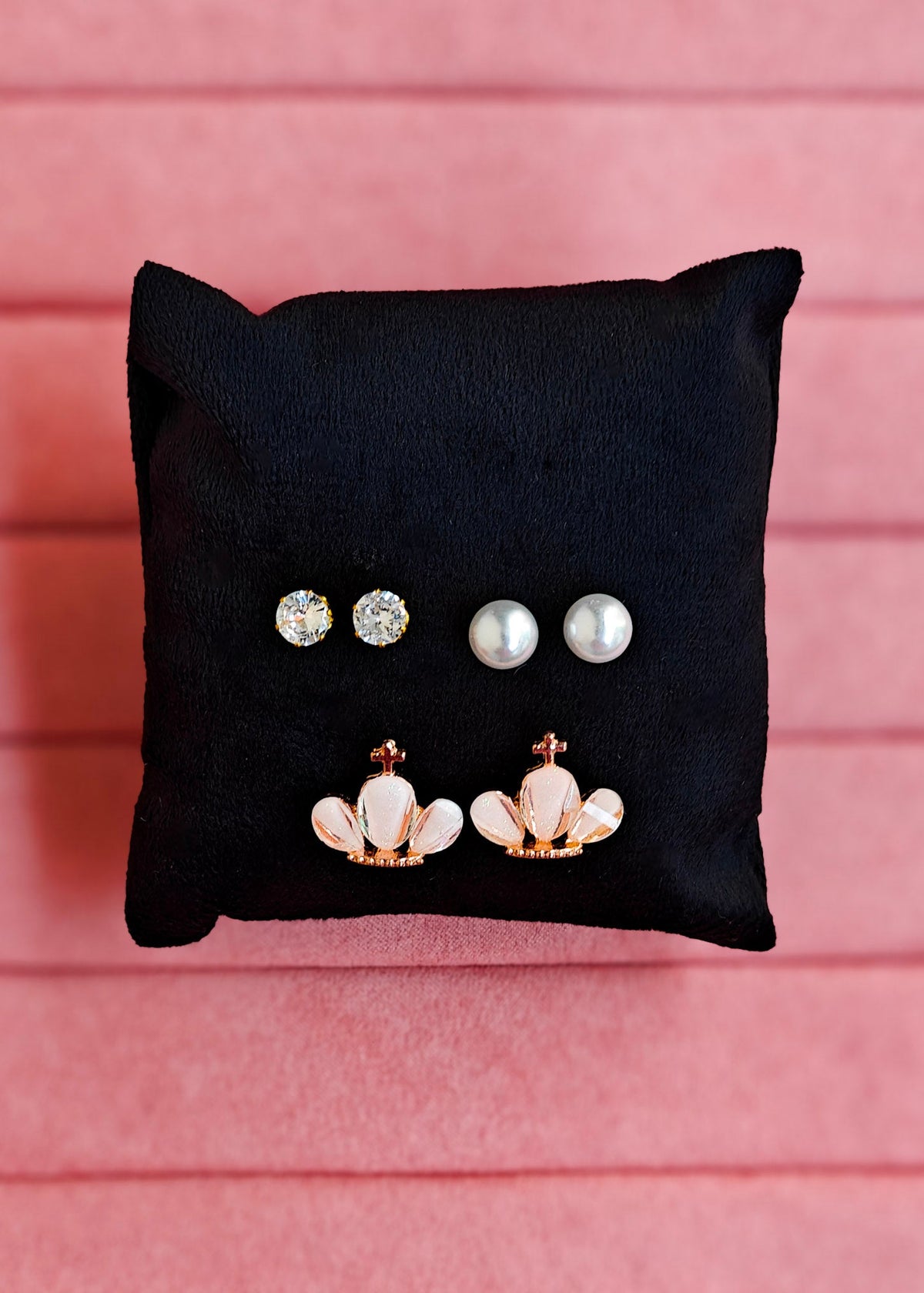 Three pairs of stud earrings displayed on a black fabric cushion against a pink paneled background: two silver-toned round stud earrings, two white pearl stud earrings, and two rose gold flower-shaped drop earrings with white inlays.