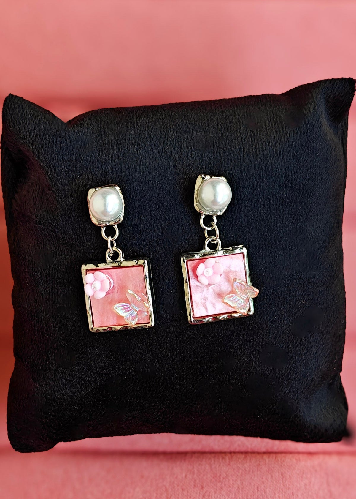 A pair of dangling earrings displayed on a black cushion against a pink background. Each earring has a round white pearl-like top, a silver-tone chain, and a square pink floral enamel or mother-of-pearl pendant with butterfly details.
