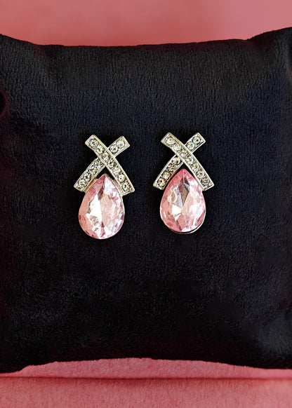 A pair of silver-toned earrings with a teardrop-shaped pink crystal drop and an X-shaped top set with small clear crystals, displayed on a black velvet cushion against a pink background.