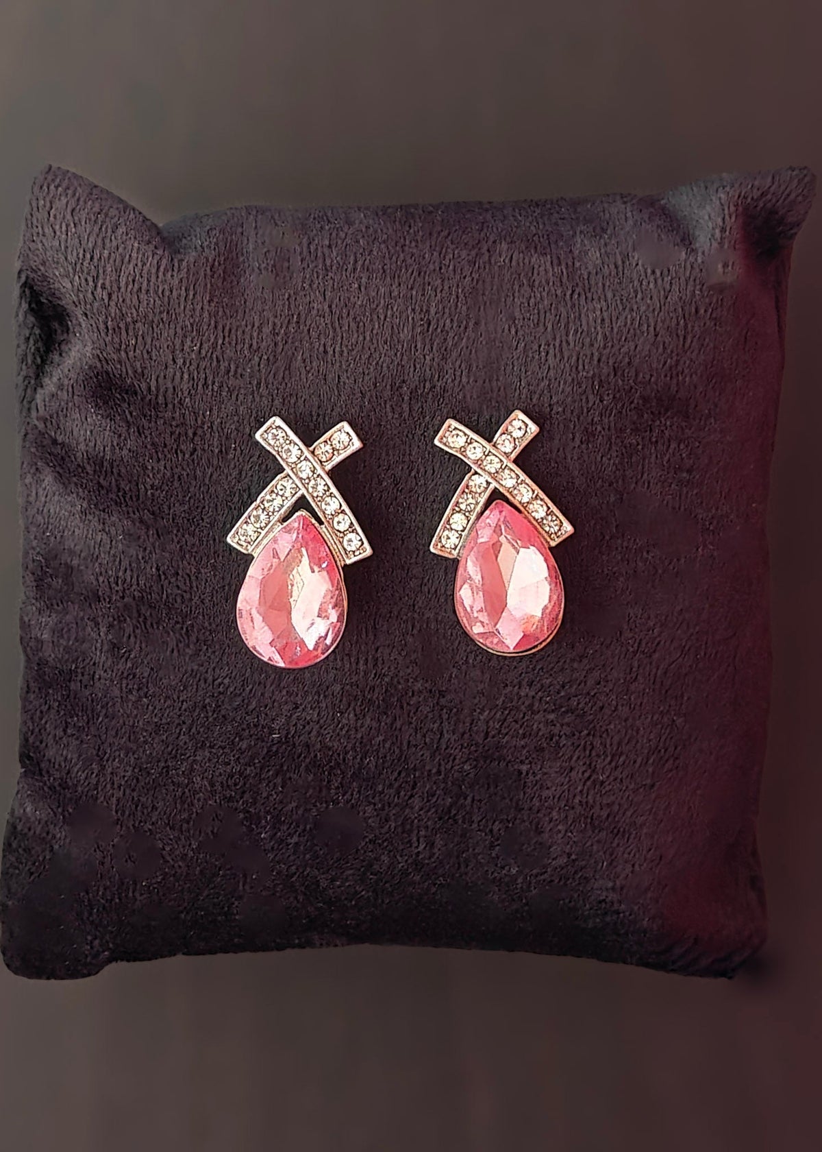 A pair of silver-toned cross stud earrings with clear rhinestones and pink teardrop gemstones, displayed on a dark brown velvet cushion against a dark background.