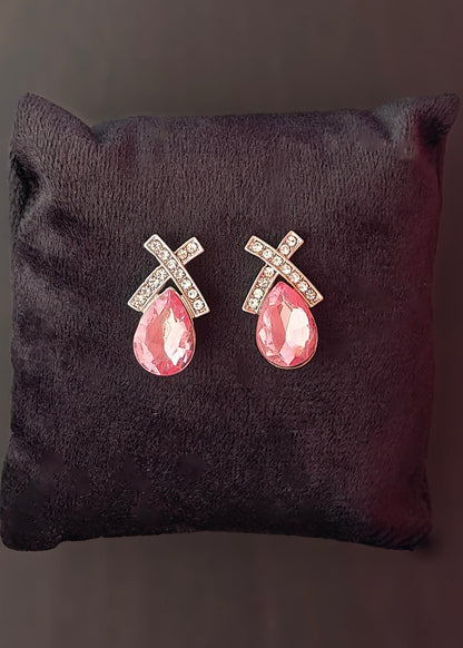 A pair of silver-toned cross stud earrings with clear rhinestones and pink teardrop gemstones, displayed on a dark brown velvet cushion against a dark background.