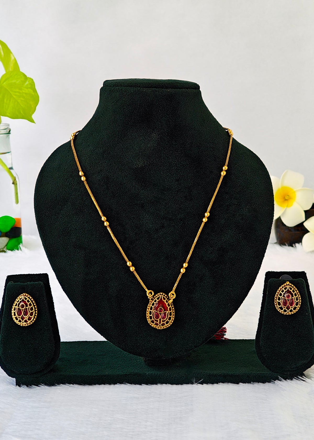 Gold necklace and matching earrings set displayed on a black velvet jewelry bust, featuring a teardrop-shaped pendant with red stone detailing.