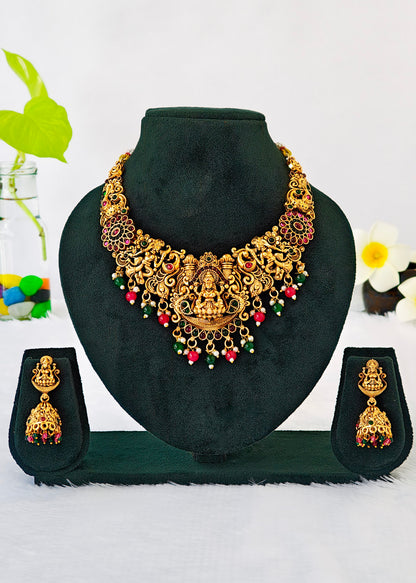 An ornate gold-toned traditional necklace set displayed on a black velvet jewelry bust, with matching drop earrings shown on small stands on both sides. The jewelry features intricate filigree detailing and red and green bead accents.