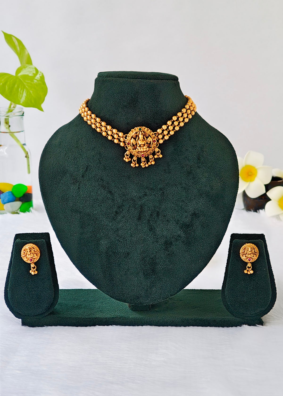 Gold-tone jewelry set displayed on a dark green velvet jewelry bust, including a necklace with a central ornate pendant and matching drop earrings, shown on a white background with decorative flowers and leaves.