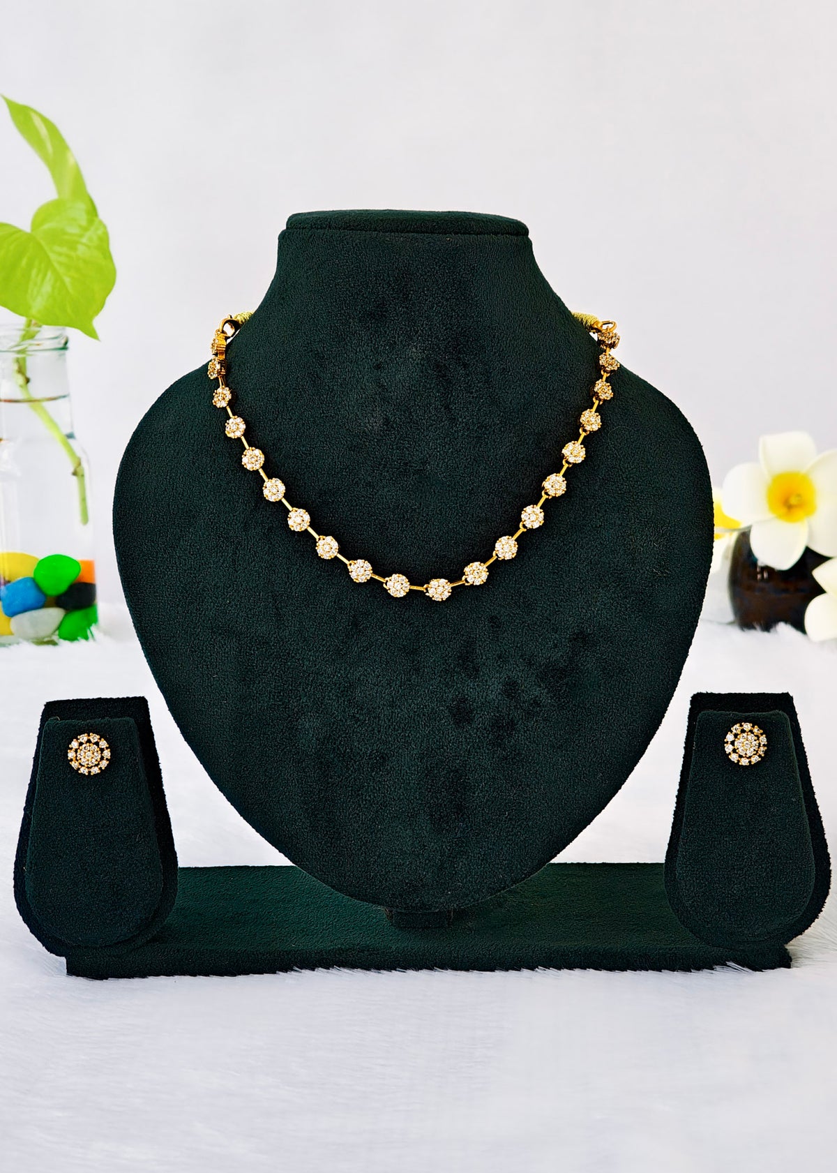 Gold beaded necklace and matching stud earrings displayed on a black velvet jewelry bust against a white background with decorative flowers and a plant.