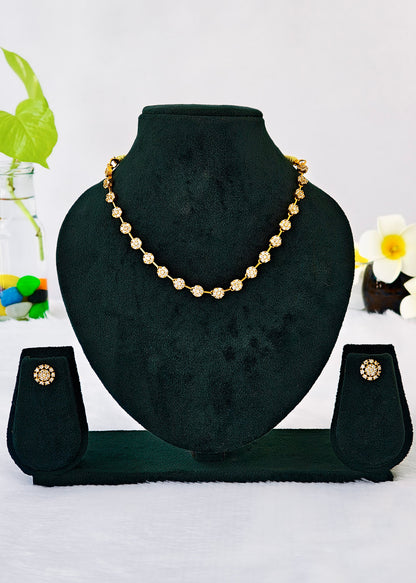 Gold beaded necklace and matching stud earrings displayed on a black velvet jewelry bust against a white background with decorative flowers and a plant.