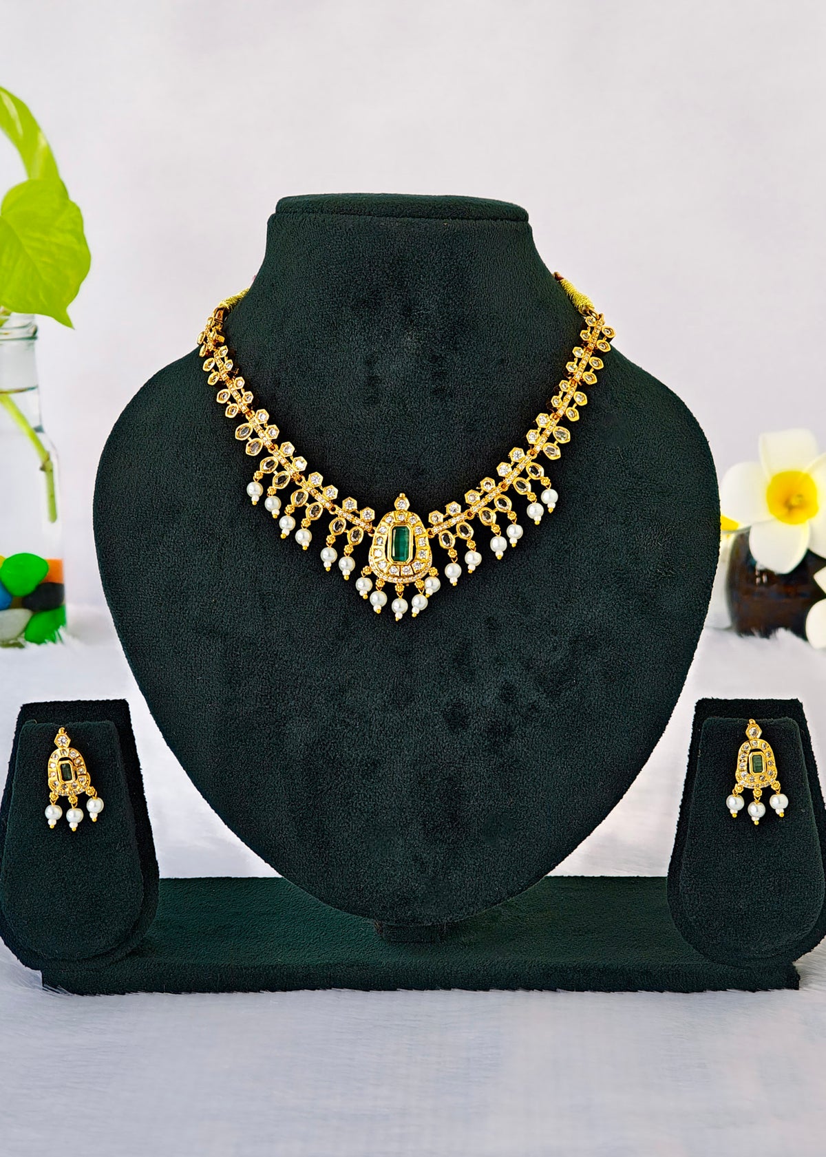 Gold-toned traditional necklace and matching earrings displayed on a black jewelry bust, with small decorative flowers in the background.