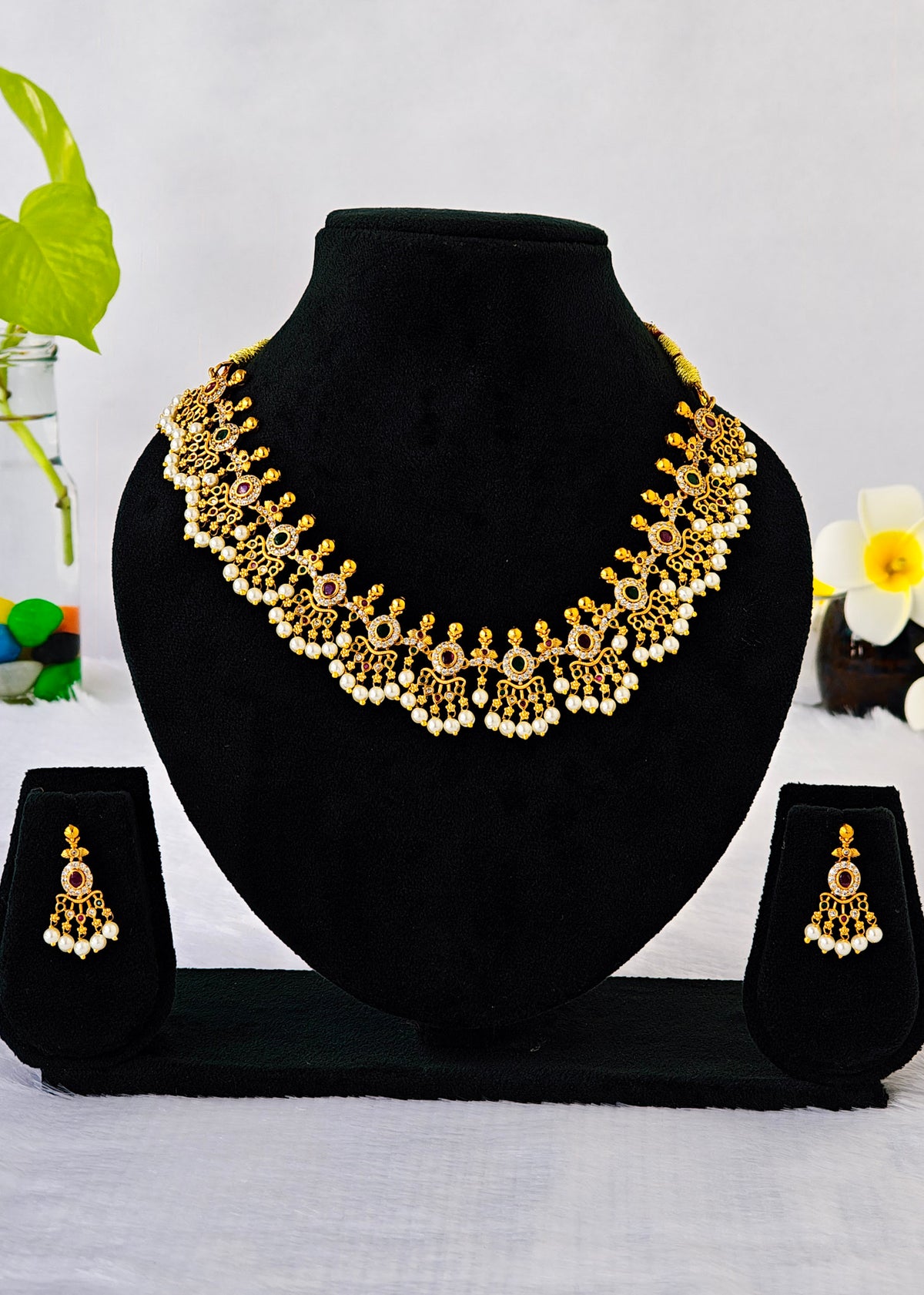 Gold-toned traditional necklace and matching dangle earrings displayed on a black velvet jewelry bust with two small matching display stands, set against a light background with small floral props.