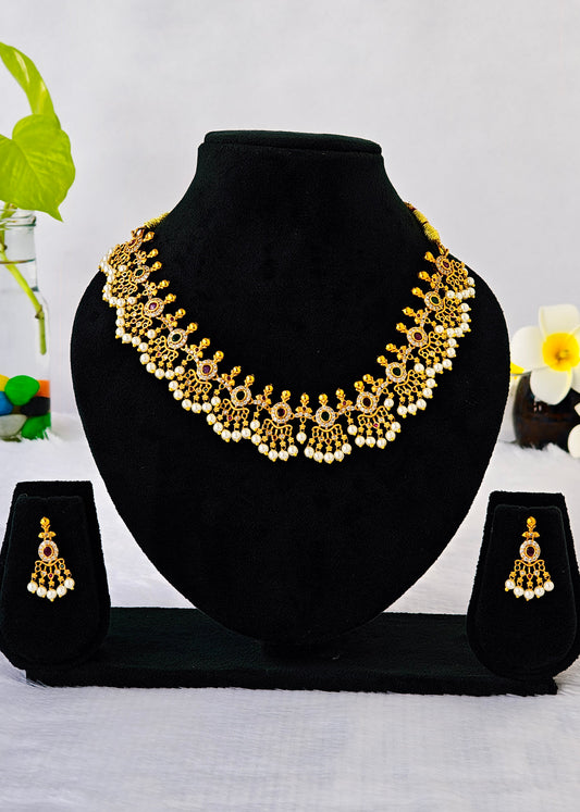 Gold-toned traditional necklace and matching dangle earrings displayed on a black velvet jewelry bust with two small matching display stands, set against a light background with small floral props.