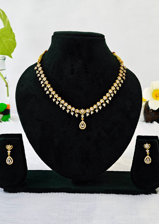 Gold-toned jewelry set displayed on a black velvet bust, including a necklace with a teardrop pendant and matching drop earrings, shown against a light background with decorative flowers.