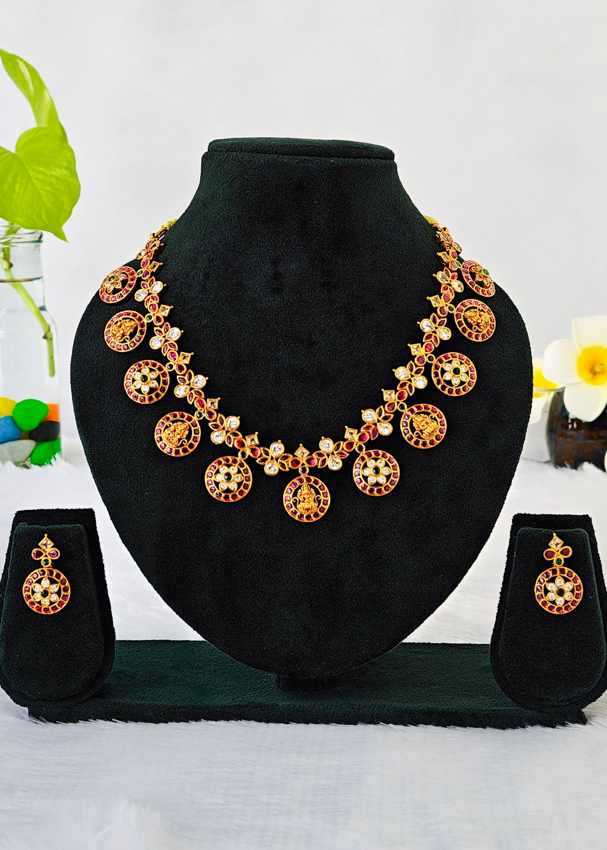Gold-toned traditional jewelry set displayed on a black velvet bust, including a necklace with circular pendant motifs, matching drop earrings, and a small stand, photographed against a light background with decorative flowers at the sides.