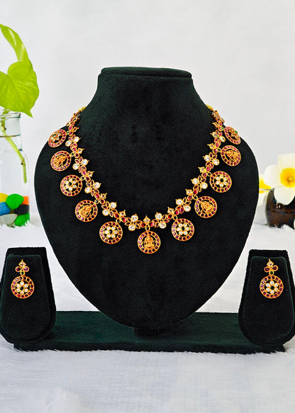 Gold-toned traditional jewelry set displayed on a black velvet bust, including a necklace with circular pendant motifs, matching drop earrings, and a small stand, photographed against a light background with decorative flowers at the sides.