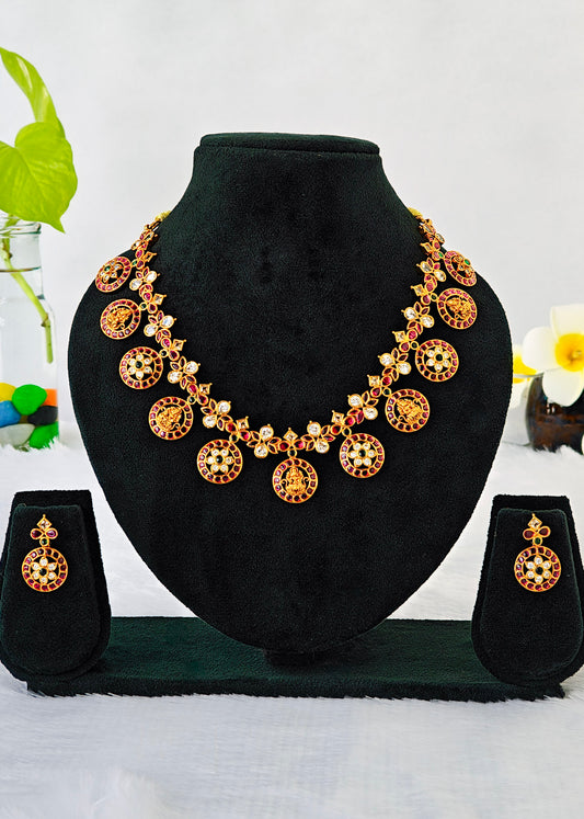 Gold-toned traditional jewelry set displayed on a black velvet bust, including a necklace with circular pendant motifs, matching drop earrings, and a small stand, photographed against a light background with decorative flowers at the sides.