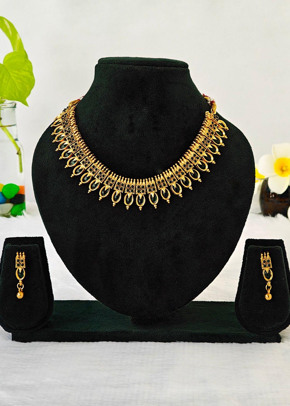 Gold-toned traditional Indian jewelry set featuring a choker necklace, matching drop earrings, and a matching ring, displayed on a black velvet jewelry bust and stand against a white background with decorative flowers.