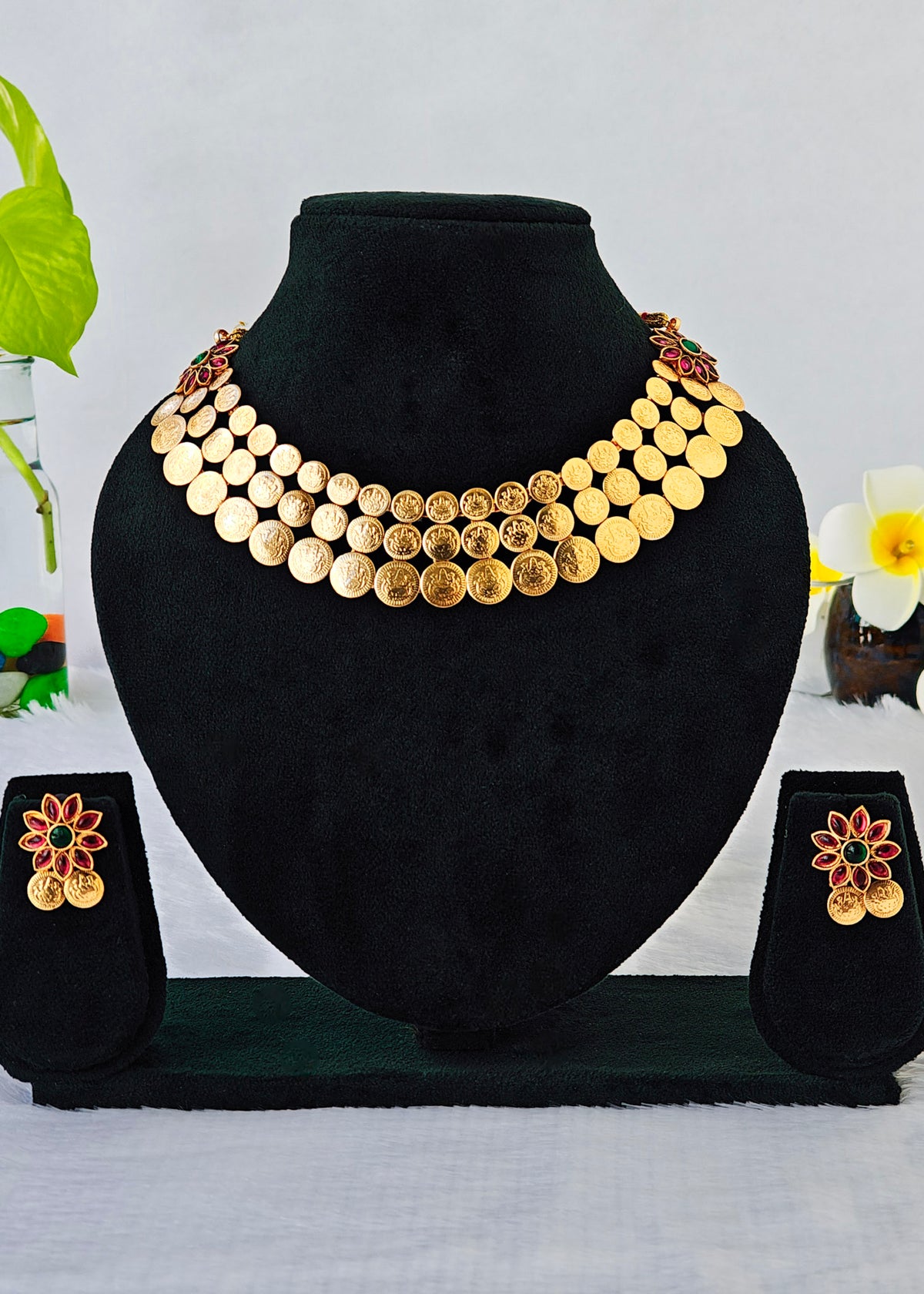 A gold coin necklace set displayed on a black velvet jewelry bust, with matching flower-shaped earrings on small display stands. The necklace features multiple rows of round gold coins with some engraved details, and the earrings have gold coin details with red and green enamel accents.
