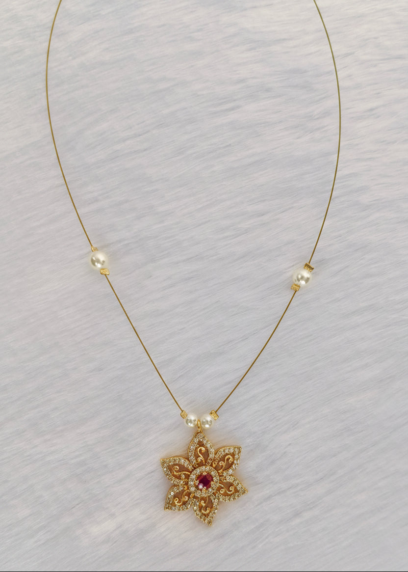A delicate gold chain necklace featuring a small gold flower pendant with a central pink gemstone and tiny clear stones, accented with small white pearl beads on a textured white background.