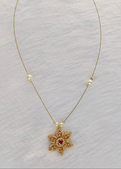 A delicate gold chain necklace featuring a small gold flower pendant with a central pink gemstone and tiny clear stones, accented with small white pearl beads on a textured white background.
