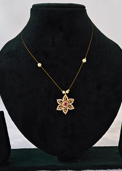 A gold necklace with a floral starburst pendant featuring a pink center, displayed on a black velvet jewelry bust. The necklace also features two small gold beads on either side of the chain, which are decorated with tiny white stones and a small pearl-like bead.