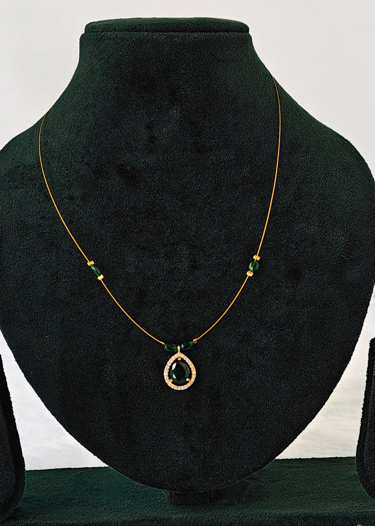 A gold chain necklace with a teardrop-shaped green gemstone pendant and small green bead accents, displayed on a black velvet jewelry bust.
