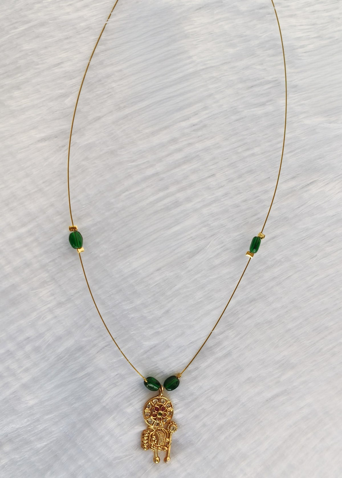 A delicate gold chain necklace featuring a small gold monkey pendant and two green oval beads spaced along the chain, displayed on a textured white fabric background.