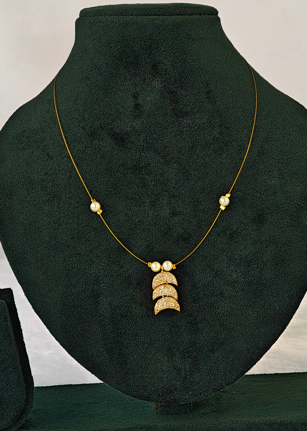 A gold necklace displayed on a black velvet jewelry bust. The necklace features a delicate gold chain with small gold bead accents on either side of the chest, and a central pendant composed of three stacked gold crescent moon shapes with small pearl-like beads at the top.