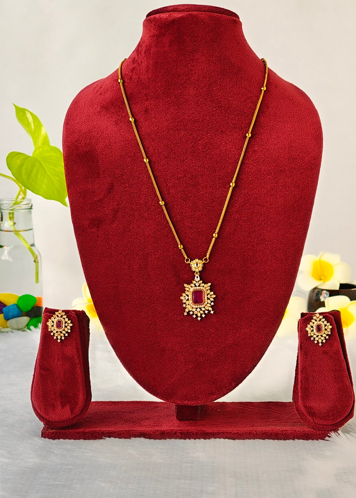 Gold-toned traditional Indian jewelry set featuring a necklace with a rectangular red stone pendant, a matching bracelet, and a pair of earrings, displayed on a red velvet jewelry bust.