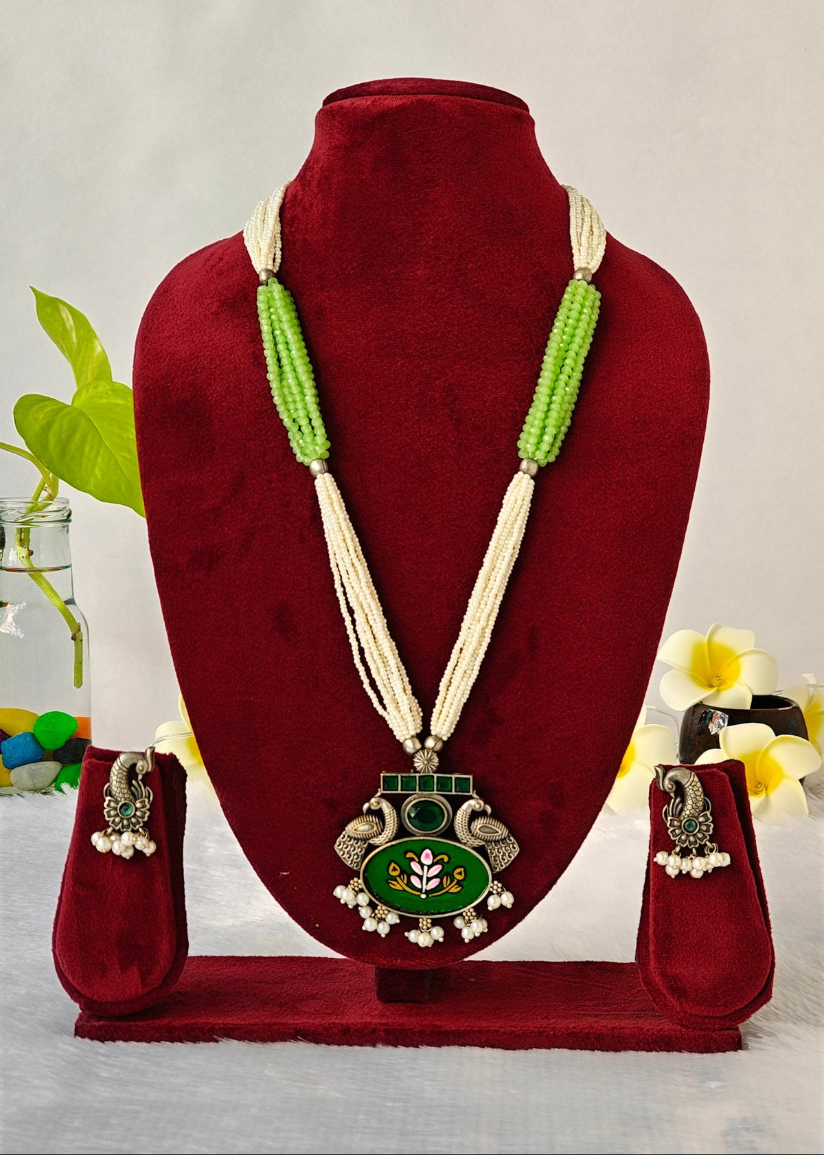 A traditional Indian jewelry set displayed on a dark red velvet mannequin bust, featuring a long necklace with a green and silver pendant, matching green beaded necklaces, and silver-toned earrings with white pearl bead drops.