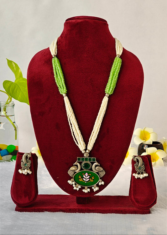 A traditional Indian jewelry set displayed on a dark red velvet mannequin bust, featuring a long necklace with a green and silver pendant, matching green beaded necklaces, and silver-toned earrings with white pearl bead drops.