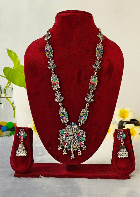 A silver-toned traditional Indian jewelry set featuring a necklace and matching earrings, embellished with colorful rhinestones, displayed on a dark red velvet jewelry stand.