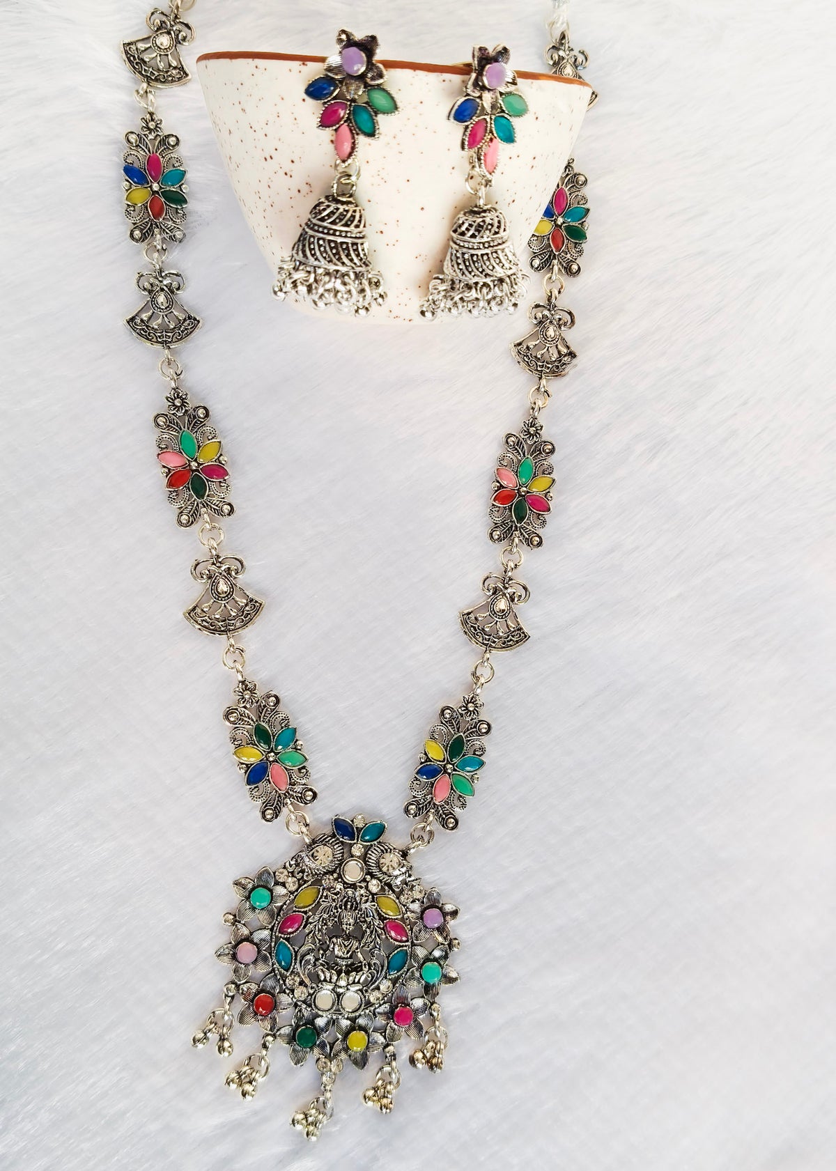 A traditional Indian jewelry set featuring a necklace, earrings, and a matching stand, decorated with silver-toned metal and vibrant multicolored stones in pink, green, blue, and yellow.