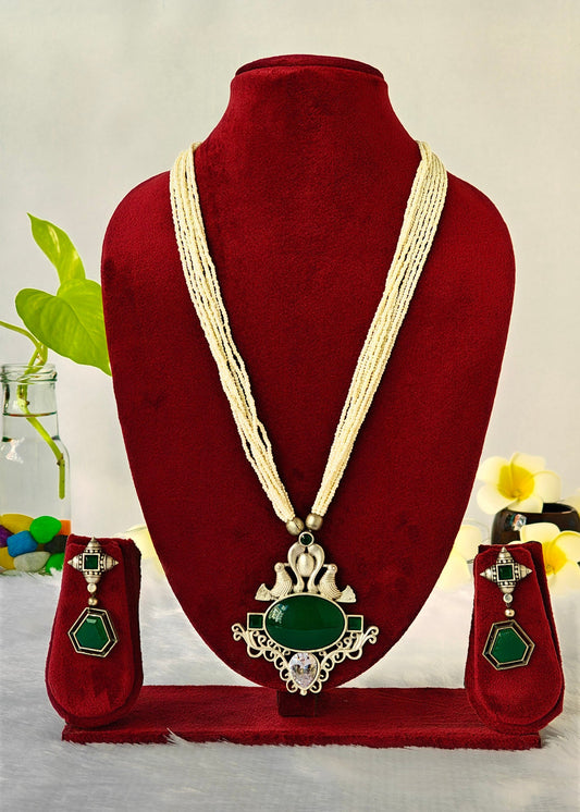A traditional Indian jewelry set displayed on a red velvet mannequin bust, featuring a multi-strand cream necklace with a large green stone pendant, matching green and silver earrings, and a red stand.