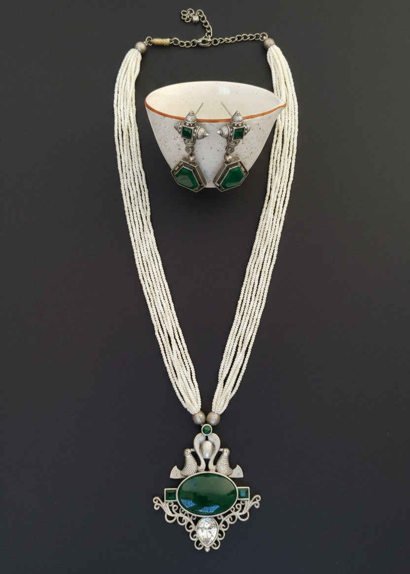 A silver-toned jewelry set featuring a multi-strand cream necklace with a large ornate pendant containing a dark green oval stone and matching green geometric drop earrings