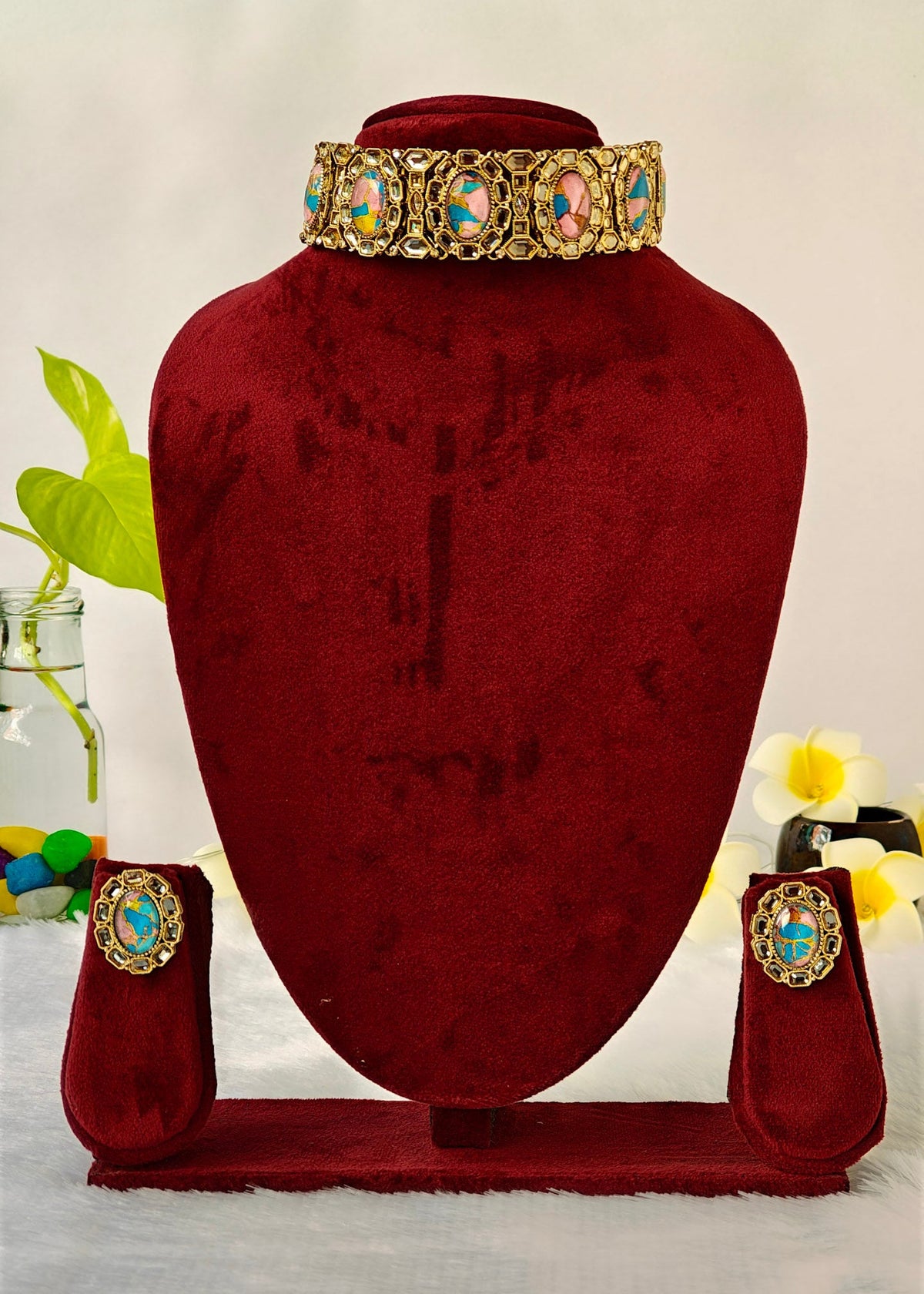 A gold-toned jewelry set displayed on a dark red velvet mannequin bust, including a wide collar necklace, matching earrings, and a small pouch, with decorative flowers and a glass vase in the background.