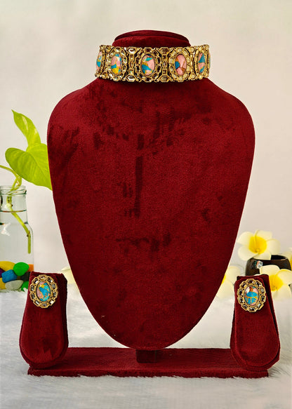 A gold-toned jewelry set displayed on a dark red velvet mannequin bust, including a wide collar necklace, matching earrings, and a small pouch, with decorative flowers and a glass vase in the background.
