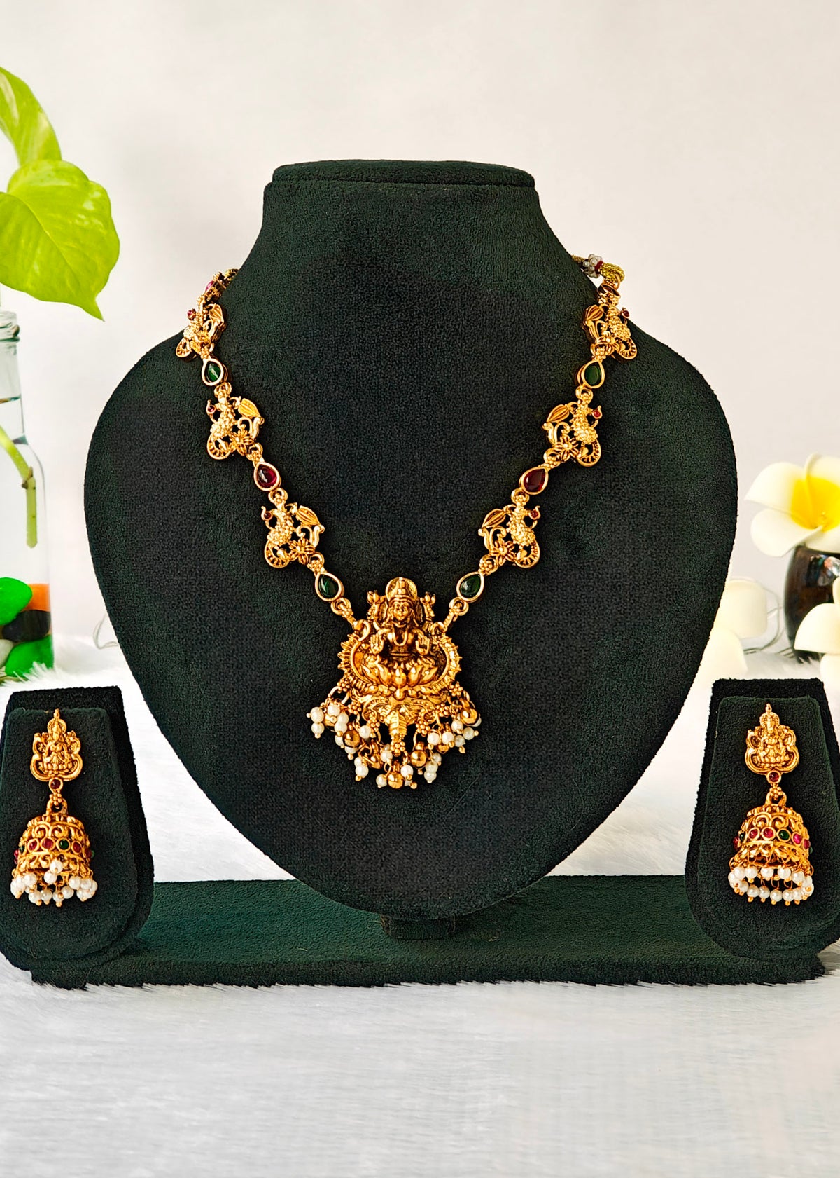Gold-toned traditional jewelry set displayed on a black velvet mannequin bust, including a necklace with a central ornate pendant and matching drop earrings, shown against a light neutral background with small decorative floral elements.