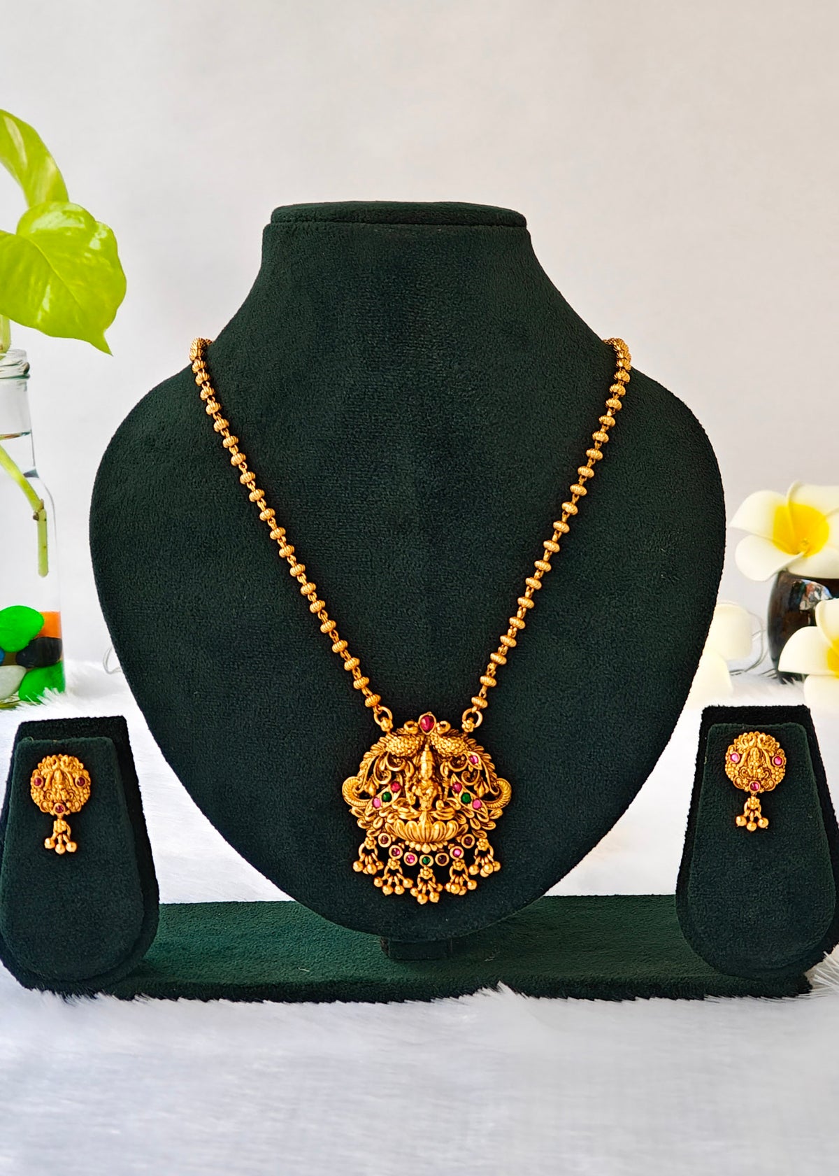 Gold necklace and matching earrings displayed on a black jewelry bust set against a light background with decorative flowers and leaves.