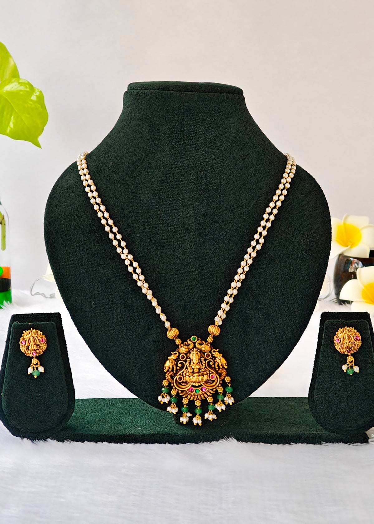 Vasudha two-strand pearl necklace with a Lakshmi and elephant pendant, green bead drops and matching earrings on a black display.