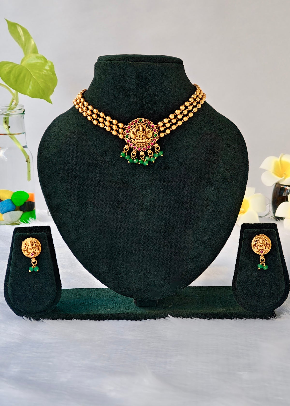Gold-tone traditional Indian jewelry set featuring a choker necklace, matching drop earrings, and a ring, displayed on a black velvet jewelry bust with a matching stand. The jewelry features gold-plated elements with green stone accents.