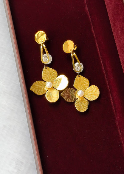 A pair of gold flower-shaped drop earrings with textured petals, small clear rhinestones, and white pearl centers, displayed on a dark red velvet jewelry box.