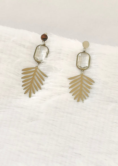 A pair of bohemian-style dangle earrings featuring gold-toned palm leaf charms, clear rectangular glass beads, and a small brown tiger's eye bead, displayed on a white textured fabric background.